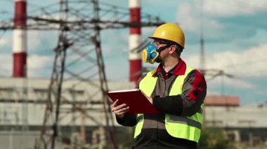 Elektrik santralindeki gaz maskeli adam iş belgelerine not alıyor. Elektrik mühendisliği uzmanı. Solunum cihazında ve sarı kaskette. Belgelerde yazıyor.