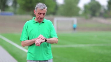 Saati not al ve koşmaya başla. Kıdemli adam futbol sahasının yakınında koşar. Yeşil tişörtlü aktif yaşlı bir adam futbol sahasının etrafında koşuyor. Fiziksel aktivite kalori yakmaya yardımcı olur.