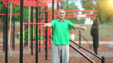Son sınıf öğrencisi spor sahasında fiziksel egzersiz yapıyor. Yeşil tişörtlü aktif yaşlı adam dışarıda spor salonunda egzersiz yapıyor. Gri saçlı sporcu yorucu bir egzersiz yapıyor, sabah güneşi arka planda.