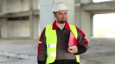 İnşaat alanındaki komik inşaatçı surat yapıyor. Başlıklı işçi proje sitesinde duruyor, kameraya bakıyor ve suratını ekşitiyor.