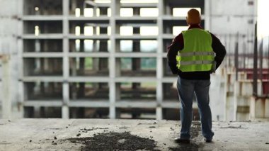 Usta inşaatçı gün batımında yeni şantiyeye bakıyor. Başlıklı işçi proje sitesinde duruyor ve yeni bina yapısına bakıyor