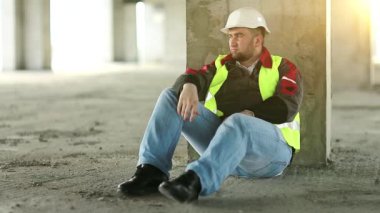 Beyaz şapkalı bir bina beton zeminde oturuyor. İşçi inşaat alanında dinleniyor, arka planda güzel bir güneş var.