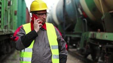 Demiryolu işçisi yük trenlerinin yanında cep telefonuyla konuşuyor. Demiryolu çalışanı demiryolu hattında çalışıyor. Cep telefonuyla konuşuyor. İşyerinin müdürü, nakliye istasyonundaki akıllı telefon aracılığıyla iletişim kuruyor.