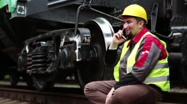 Neşeli demiryolu işçisi cep telefonuyla konuşuyor. Demiryolu çalışanı iş kıyafetleri içinde yük vagonunun yanında oturur ve cep telefonuyla konuşur.
