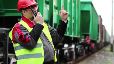 Demiryolu işçisi, yük trenlerinin yanında taşınabilir radyo vericisi var. Demiryolu çalışanı, nakliye istasyonunun yanındaki demiryolu hattında portatif radyo vericisi aracılığıyla konuşuyor. İşçi radyo istasyonu aracılığıyla iletişim kuruyor.
