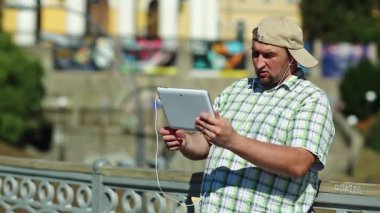 Kareli gömlekli ve beysbol şapkalı şişman çocuk elinde kulaklıkla beyaz tablet tablet tutuyor. Adam tablet bilgisayarla konuşuyor. Tabletli adam video kaset aracılığıyla iletişim kuruyor.