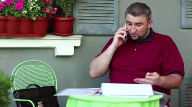 Duygusal iş adamı, kafede akıllı telefondan konuşur ve iş belgeleriyle çalışır. Adam yaz restoranında masada oturuyor ve cep telefonuyla konuşuyor, tablet bilgisayar masanın üstünde.