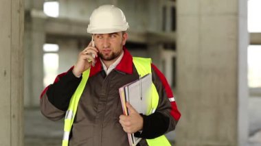 Yeşil yelek giyen inşaatçı proje alanında telefonla konuşuyor. Beyaz kasklı işçiler inşaat alanında akıllı telefondan konuşuyor.