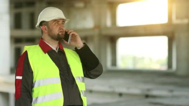 İnşaatçı inşaat alanında cep telefonuyla konuşuyor. Yeşil yelek ve beyaz şapka giyen işçiler proje sitesindeki akıllı telefondan konuşuyor