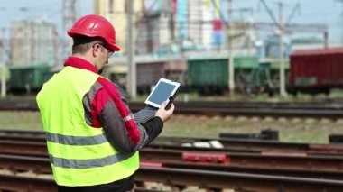 Yük treni terminalinde bilgisayarlı bir demiryolu işçisi. Demiryolu çalışanı tabletine not alıyor. Üniformalı ve kırmızı şapkalı demiryolu işçisi tablet bilgisayarla çalışıyor.