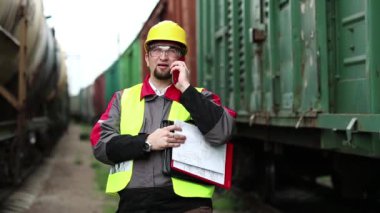 Demiryolu işçisi yük trenlerinin yanında cep telefonuyla konuşuyor. Demiryolu çalışanı yük trenlerinin arasında duruyor ve cep telefonuyla konuşuyor. İşletme müdürü, nakliye istasyonundaki akıllı telefon aracılığıyla iletişim kuruyor.