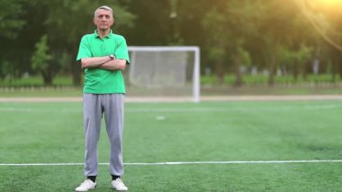 Futbol antrenörü futbol sahasında, beden eğitimi öğretmeni. Yeşil tişörtlü adam futbol sahasında duruyor ve kameraya bakıyor.