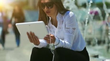 İş kadını fıskiyelerin yanında oturur tabletle iletişim kurar ve kahve içer. Beyaz tablet bilgisayarlı gözlüklü çekici bir kadın görüntülü sohbet yoluyla iletişim kuruyor.