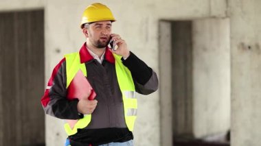 İnşaat alanındaki inşaat konuşmalarında sarı sapkalı iş belgeleriyle çalışan ustabaşı. İnşaatçı proje sitesindeki cep telefonundan konuşuyor