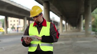 Üstgeçidin altında tablet bilgisayarı olan bir işçi. Tamirat müdürü inşaat sahasında çalışıyor. Ana yolun altında. Bilgisayarına veri giriyor.