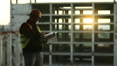 Proje sahasında çalışma belgelerine sahip bir inşaatçı. Usta müteahhit iş belgelerine notlar alır. Bina müfettişi belgelerine arka planda güzel bir güneş olduğunu yazmış.