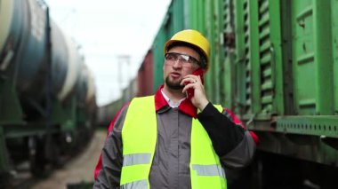 Demiryolu işçisi tren yakınlarında cep telefonuyla konuşuyor. Demiryolu çalışanı demiryolu hattında çalışıyor. Cep telefonuyla konuşuyor. İşyerinin müdürü, nakliye istasyonundaki akıllı telefon aracılığıyla iletişim kuruyor.