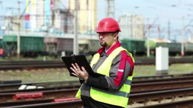 Yük treni terminalinde bilgisayarlı bir demiryolu işçisi. Demiryolu çalışanı tabletine not alıyor. Üniformalı ve kırmızı şapkalı demiryolu işçisi tablet bilgisayarla çalışıyor.