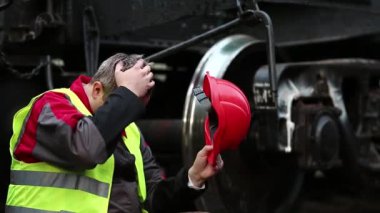 Depresyondaki kırmızı şapkalı demiryolu işçisi trajik olaylardan sonra yük vagonunun yanında oturur. Demiryolu raylarında stresli demiryolu işçisi, demiryolu hattında büyük metal tekerleğin yanında oturuyor.