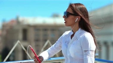 Genç bir kadın müzik dinler ve şarkı söyler. Uzun saçlı, güneş gözlüklü, kırmızı akıllı telefonlu güzel kız şehirde müzik dinliyor.