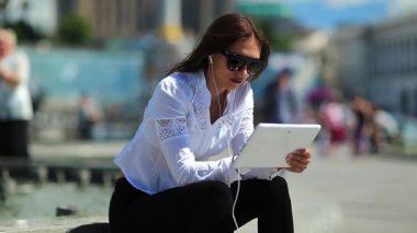 Kadın çeşmelerin yanında oturuyor ve tablet bilgisayar kullanıyor. Elinde beyaz tablet bilgisayarla gözlüklü çekici bir kadın.