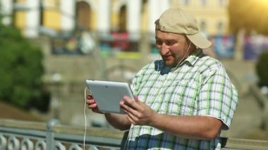 Kareli gömlekli ve beysbol şapkalı şişman çocuk elinde kulaklıkla beyaz tablet tablet tutuyor. Adam tablet bilgisayarla iletişim kuruyor. Tabletli adam video kaset aracılığıyla iletişim kuruyor.