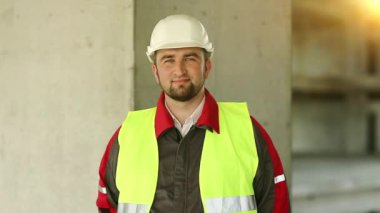 Yeni inşaat alanındaki usta inşaatçı kameraya bakıyor. Başlıklı işçi proje alanında duruyor, arka planda güzel bir güneş var.