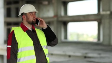 Yeşil yelekli inşaatçı proje alanında cep telefonuyla konuşuyor. Beyaz kasklı mühendis inşaat alanında akıllı telefondan konuşuyor.