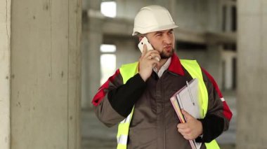 Beyaz kasklı mühendis inşaat alanındaki akıllı telefondan konuşuyor. Yeşil yelek giyen inşaatçı proje alanında telefonla konuşuyor. İşçiler el ele tutuşur. Gazeteler ve cep telefonları çalışır.