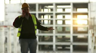 İşçi inşaat alanında akıllı telefondan konuşur. Projenin mimarı cep telefonuyla konuşuyor. Arka planda güzel bir günbatımı var.