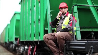 Demiryolu işçisi yeşil vagonda oturur ve cep telefonuyla konuşur. Demiryolu çalışanı demiryolu hattında çalışıyor. Cep telefonuyla konuşuyor. İşyerinin müdürü, nakliye istasyonundaki akıllı telefon aracılığıyla iletişim kuruyor.