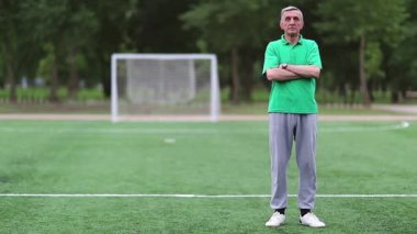 Futbol antrenörü futbol sahasında, beden eğitimi öğretmeni. Yeşil tişörtlü adam futbol sahasında duruyor ve kameraya bakıyor.
