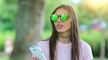 Çekici kız şehir parkında durur, müzik dinler ve dans eder. Genç kadın müzik dinler ve dans eder. Yeşil gözlüklü, akıllı telefon ve kulaklıklı güzel bir kadın doğada müzik dinliyor.
