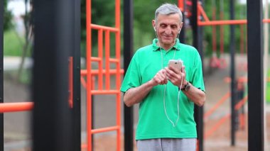 Yaşlı sporcu spor alanında akıllı telefon kullanır. Kıdemli adam cep telefonuyla çalışıyor. Gri saçlı aktif atlet açık hava spor salonunun yanında duruyor ve cep telefonunu tutuyor.