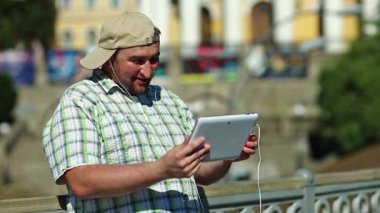 Kareli gömlekli ve beysbol şapkalı şişman çocuk elinde kulaklıkla beyaz tablet tablet tutuyor. Adam tablet bilgisayarla iletişim kuruyor. Tabletli adam video kaset aracılığıyla iletişim kuruyor.