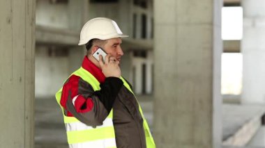 Beyaz kasklı mühendis inşaat alanındaki akıllı telefondan konuşuyor. Yeşil yelek giyen inşaatçı proje sitesinde telefonla konuşuyor