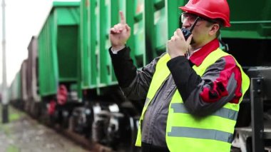 Demiryolu işçisi, yük trenlerinin yanında taşınabilir radyo vericisi var. Demiryolu çalışanı, nakliye istasyonunun yanındaki demiryolu hattında portatif radyo vericisi aracılığıyla konuşuyor. İşçi radyo istasyonu aracılığıyla iletişim kuruyor.