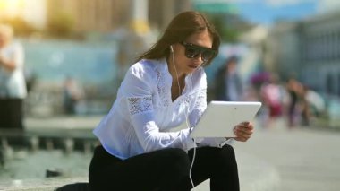 Kadın sokakta oturuyor ve tablet kullanıyor. Elinde beyaz tablet bilgisayarla gözlüklü çekici bir kadın.