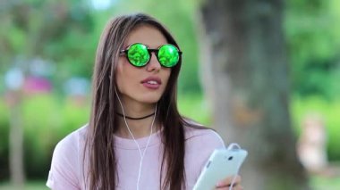 Genç kadın müzik dinler ve dans eder. Çekici kız şehir parkında durur, müzik dinler ve dans eder. Yeşil gözlüklü, akıllı telefon ve kulaklıklı güzel bir kadın doğada müzik dinliyor.