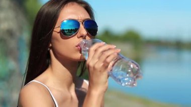 Güneş gözlüklü çekici kadın maden suyu içiyor. Bardaklı güzel kız şişeden temiz su içiyor. Kız plastik su şişesini açar ve soda içer.