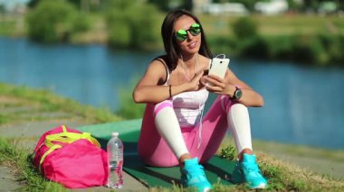 Güzel sporcu kadın göl kenarında oturuyor ve cep telefonu kullanıyor. Spor kıyafetleri ve pembe spor çantalı güneş gözlüklü güzel bir kız göletin yanında oturuyor ve cep telefonu kullanıyor. Cep telefonlu spor giyimli bir kadın.