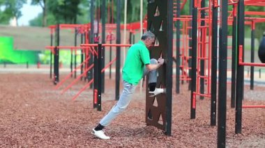Açık hava spor salonunda yeşil tişörtlü aktif yaşlı bir sporcu. Yaşlı, gri saçlı sporcu yorucu bir egzersiz yapıyor. Son sınıf öğrencisi spor sahasında fiziksel egzersiz yapıyor.