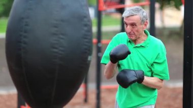Kıdemli boksör kum torbasıyla boks yapıyor. Bir boks kulübünde boks eldiveni eğitimi almış yaşlı bir boksör. Boksör kum torbasına vurur. Yeşil tişörtlü gri saçlı adam spor sahasında antrenman yapıyor.