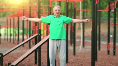 Yeşil tişörtlü aktif yaşlı adam dışarıda spor salonunda egzersiz yapıyor. Son sınıf öğrencisi spor sahasında fiziksel egzersiz yapıyor. Gri saçlı sporcu yorucu bir egzersiz yapıyor, sabah güneşi arka planda.