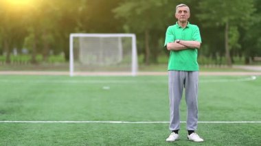 Futbol antrenörü futbol sahasında duruyor. Yeşil tişörtlü son sınıf öğrencisi futbol sahasında duruyor ve kameraya, futbol kapısına ve sabah güneşine bakıyor. Beden eğitimi öğretmeni