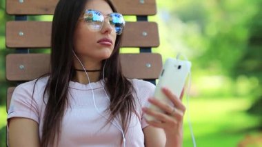 Genç ve güzel bir kız bankta oturur ve onun akıllı telefonunda müzik dinler. Akıllı telefon ve kulaklıklı çekici bir kadın müzik dinliyor.