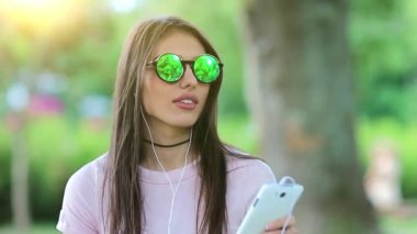 Genç ve güzel bir kız el ele tutuşup beyaz bir telefondan müzik dinliyor ve dans ediyor. Güneş gözlüklü çekici bir kadın. Kulaklıklı. Doğada müzik dinliyor.
