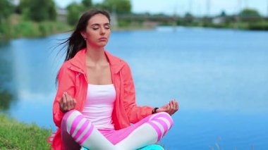 Spor kıyafetli kız gölün kenarında oturup meditasyon yapıyor. Güzel bir kadın mavi göletin yanında meditasyon yapıyor. Meditasyon - bireyin zihni çalıştırdığı, eğittiği ya da bir bilinç biçimini tetiklediği uygulama