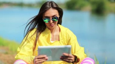 Güzel kız gölün yanında oturuyor ve tablet bilgisayarla konuşuyor. Sarı spor ceketli sporcu kadın göletin yanında oturuyor ve tableti elinde tutuyor. Spor kıyafetli bir kadın tablet bilgisayarla Skype üzerinden iletişim kuruyor.