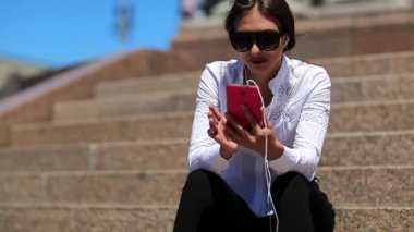 Genç bir kadın merdivenlerde oturur ve müzik dinler. Uzun saçlı, güneş gözlüklü, akıllı telefonlu güzel kız şehir merkezinde müzik dinliyor.